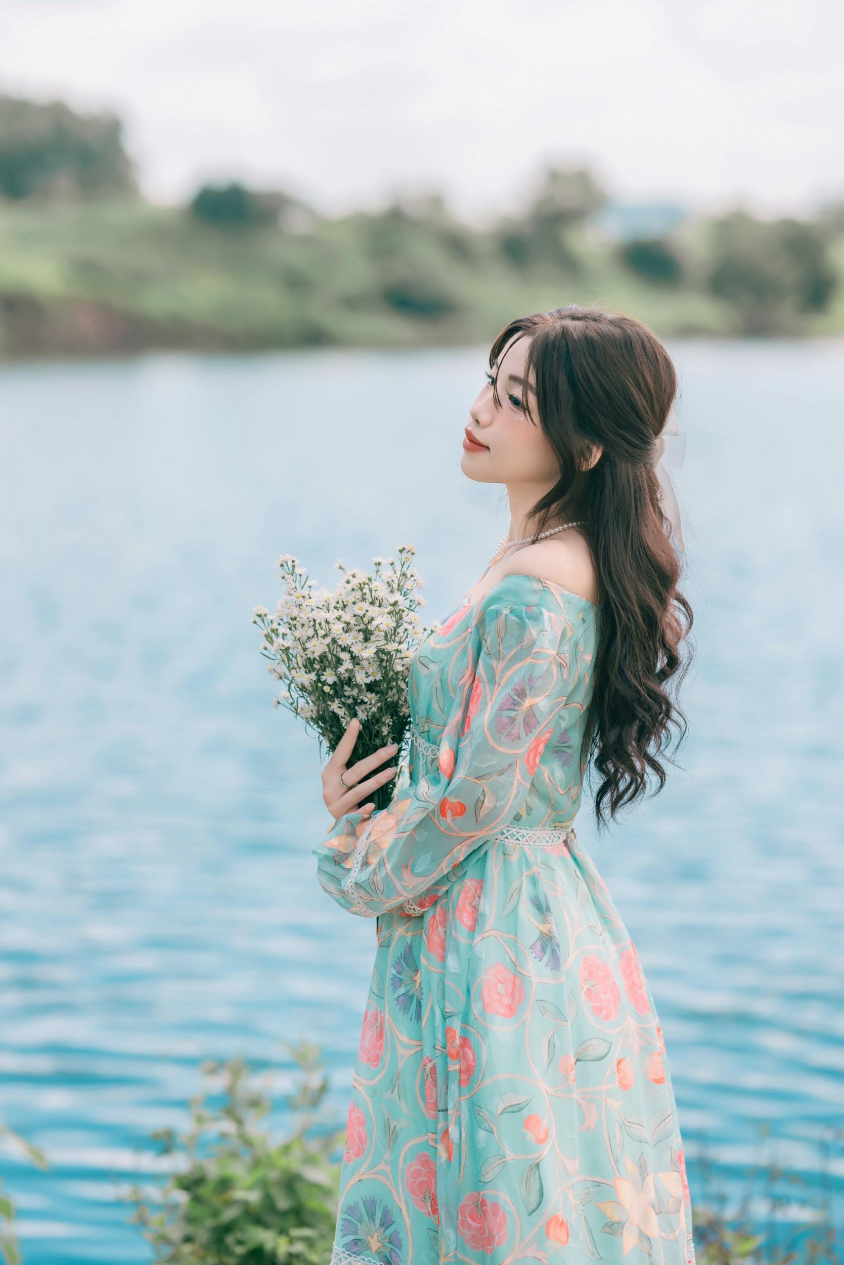 Woman in Floral Dress Holding Flowers by a Lake