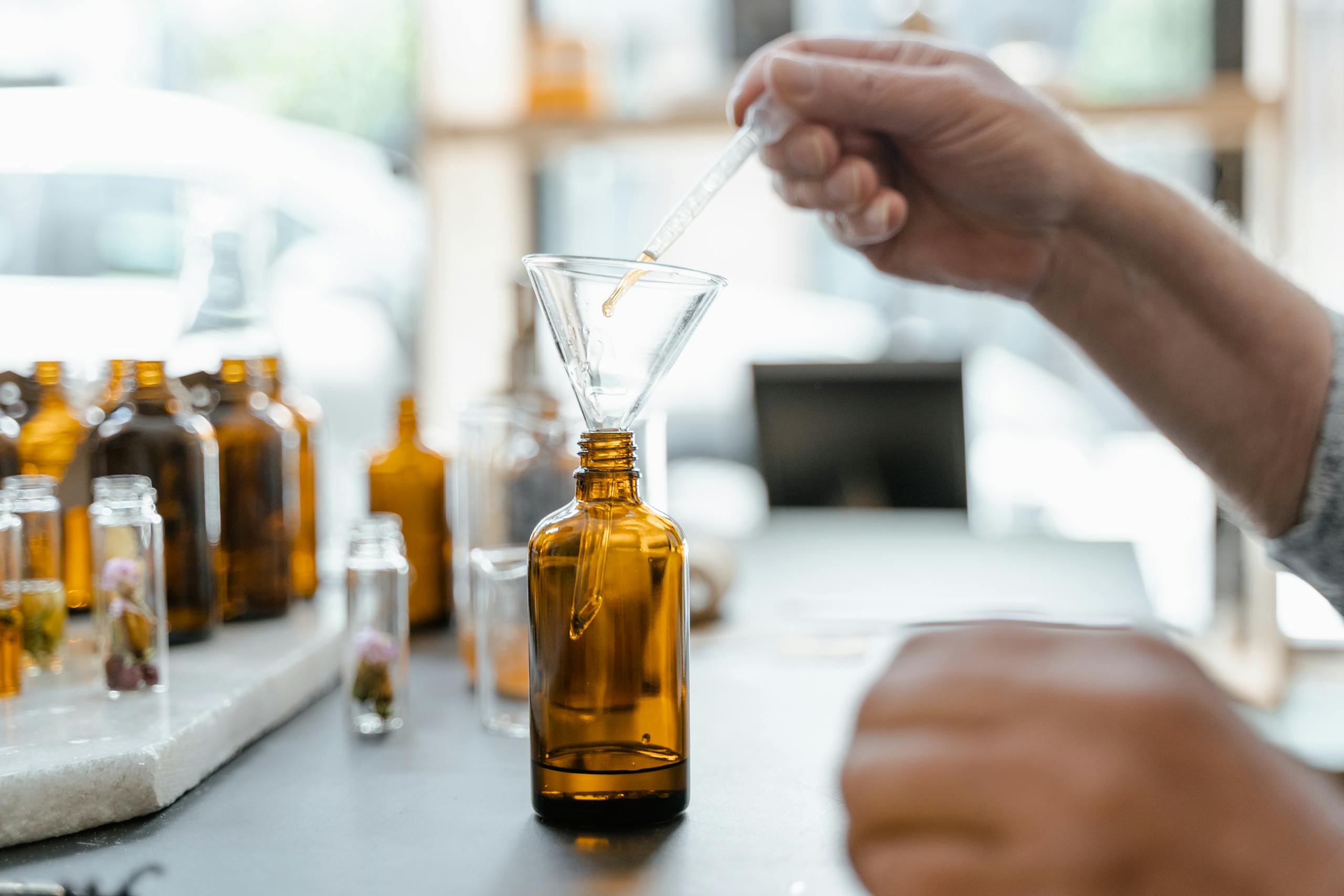 Hand pouring liquid into amber bottle using funnel, surrounded by glass jars indoors.